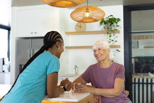 Senior and African American women sitting and leaning, holding hands at kitchen island with glass
