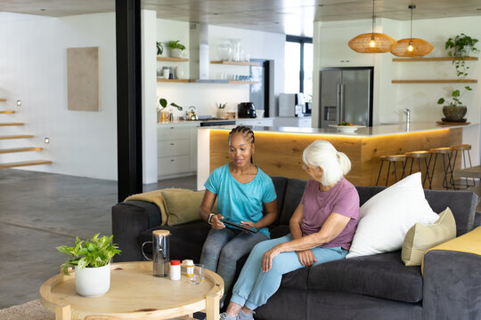 Senior woman and African American woman sitting on dark gray sectional sofa using tablet and stylus