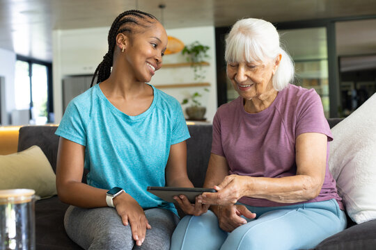 African American woman and senior woman sitting on sofa holding tablet and smiling at home