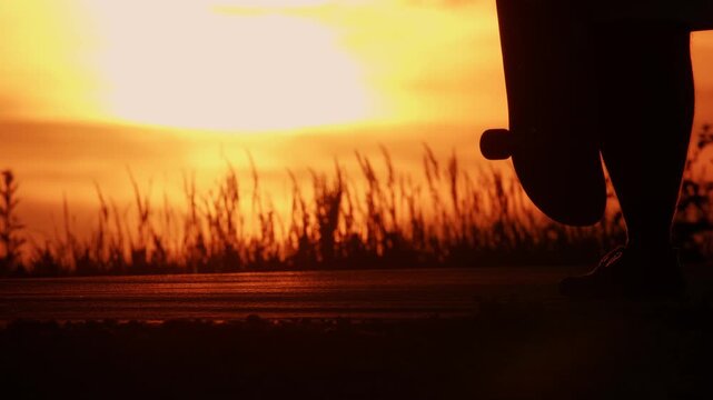 A silhouette of a man against an orange sunset and a big bright sun, close-up of his legs and skateboard, the man kicks the skateboard and catches it, throws it onto the road, jumps on the skateboard 