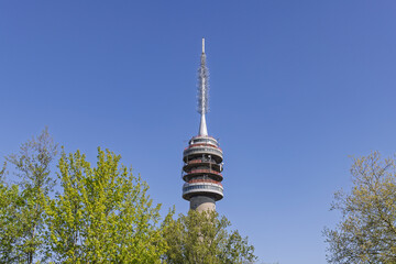 Fototapeta premium Goes, Netherlands: Tall TV and telecommunications tower standing high above lush green tree foliage against a vibrant blue sky on a clear sunny day.