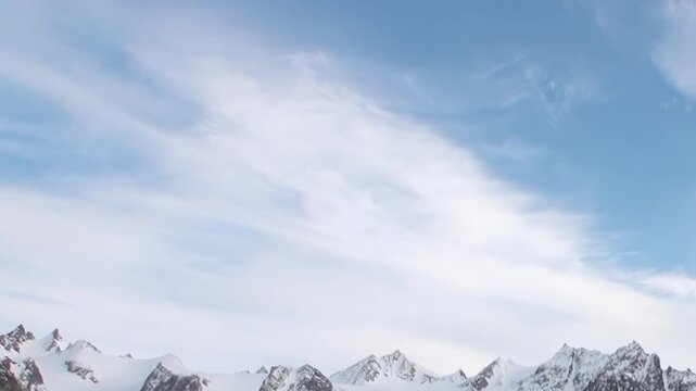 Witness grand beauty of Arctic glacier at midday. Majestic snow-capped mountains stand tall beneath expansive blue sky, painted with ethereal wisps of white clouds, revealing untouched polar realm.