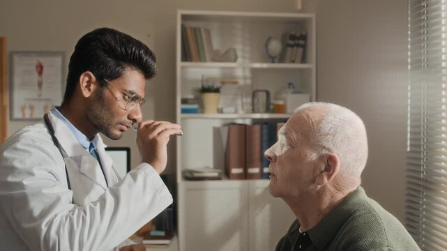 South Asian doctor examining senior Caucasian patient eyes with penlight in clinic. Video ideal for healthcare services, geriatric care marketing, and medical insurance commercials