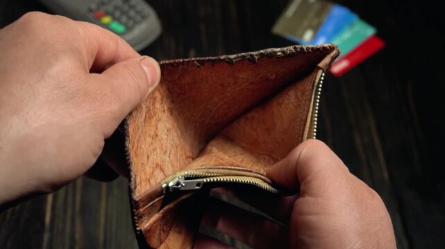 Hands of a person opening a brown leather wallet to reveal its interior, showcasing the zipper and empty compartments, with credit cards and a payment terminal visible in the background