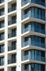 Obraz premium Modern residential building facade with balconies and large windows. White exterior walls feature glass railings and dark frames. New apartment complex under construction or recently completed.