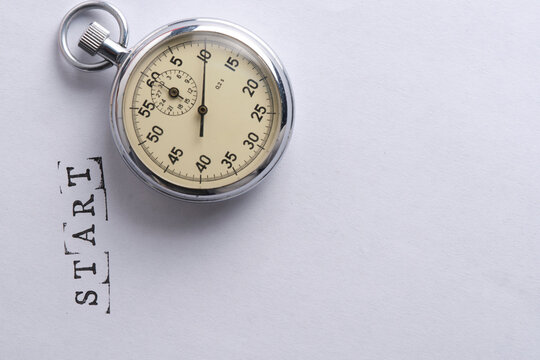 Overhead view mechanical stopwatch placed above word START stamped on paper surface.
