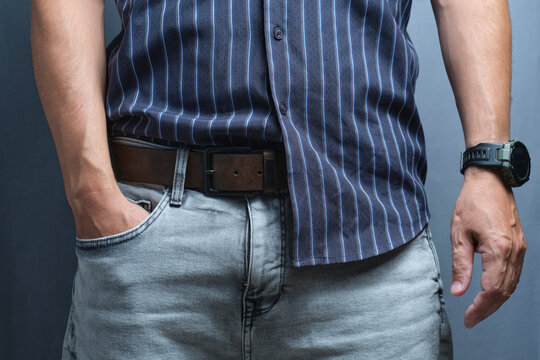 Casual man wearing grey jeans, brown leather belt, striped shirt, and watch with hand in pocket.