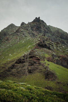 Majestic rugged mountain with vibrant green slopes and dark rock formations under a soft, cloudy sky. A breathtaking natural landscape showcasing wild beauty and untouched nature in a remote setting.