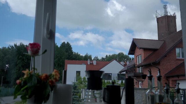 Clouds moving over houses outside window, with flowers and kitchen items near the glass. Concept of weather change and neighborhood view
