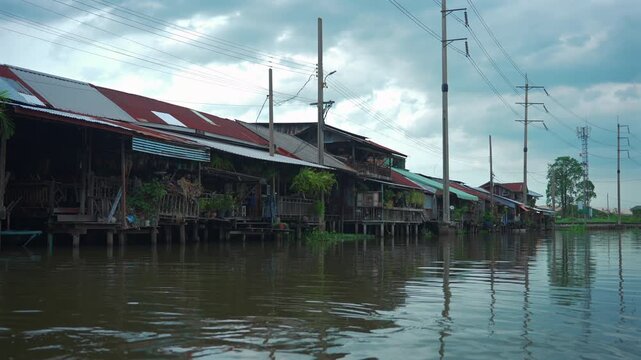 Traditional stilt houses along canal in Thailand with wooden waterfront homes, Residential canal village with riverside architecture, water reflections and power lines 4k