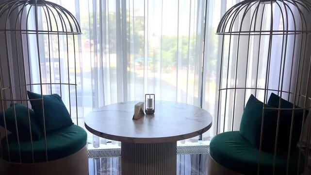 The camera slides smoothly to the right, revealing an elegant interior with a round table and unique cage chairs with turquoise cushions against the backdrop of a bright window in a cozy cafe. High