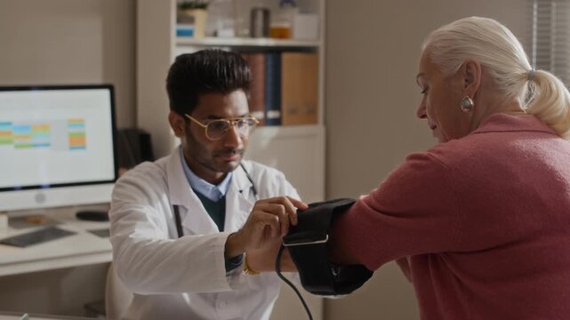 Indian male doctor putting blood pressure cuff on senior Caucasian woman arm in clinic. Perfect for healthcare services, geriatric care, and medical checkup promotional videos