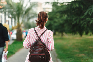 Obraz premium Young woman with a backpack walks through a green park from behind, wearing a striped shirt, capturing a casual solo moment of everyday exploration.