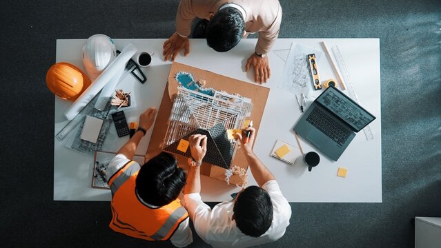 Top aerial view of architect engineer team stick sticky notes on house model while working together to design building construction at table with laptop, safety helmet and project plan. Alimentation.