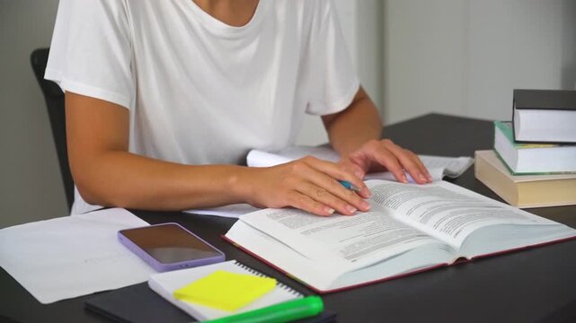 Student is reading a book and taking handwritten notes in notebook at desk with books and studies