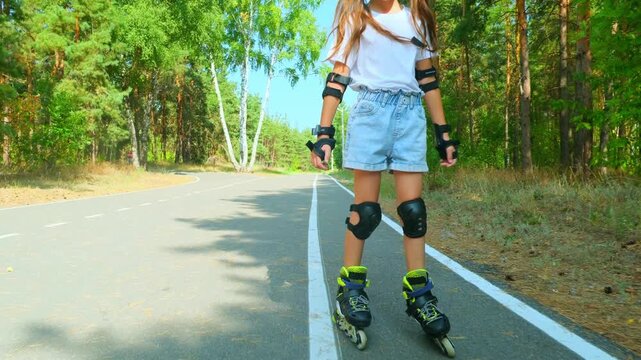 Child girl in casual clothes roller skating on road in forest on sunny summer day.