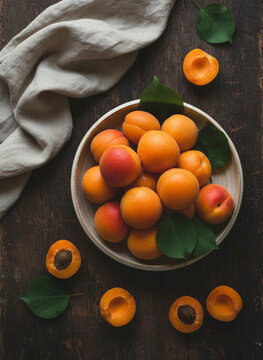 Top view of fresh ripe apricots whole and halved in a ceramic bowl on a rustic wooden table with green leaves