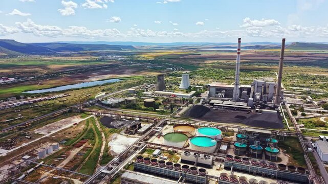 Machines and structures are seen in a large power plant near a river. Water tanks hold different colors of water. Mountains rise in the distance under a blue sky.