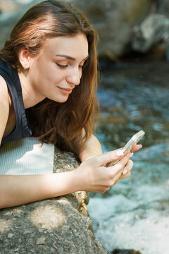 Young woman smiling while using her phone by mountain stream. Pleasant online communication, good news, outdoor lifestyle, staying connected in nature and modern digital balance