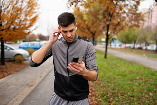 Athletic man in tracksuit standing on a park path, adjusting earphone and looking at mobile phone screen to choose playlist or track fitness data on an autumn morning.