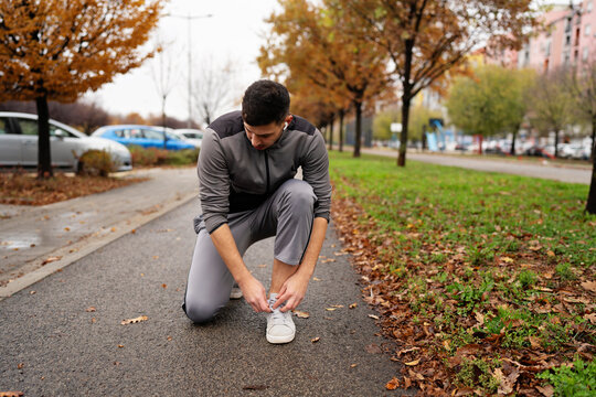 A male runner in sportswear kneels on a sidewalk in a park during autumn to fasten his sneakers before starting an outdoor workout session.