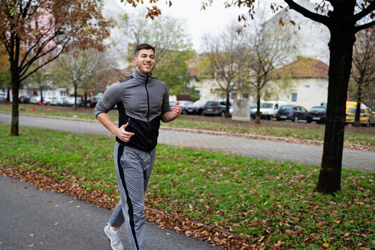 Smiling man in tracksuit enjoying a morning run through a residential park sidewalk surrounded by fall leaves, wearing wireless earbuds.