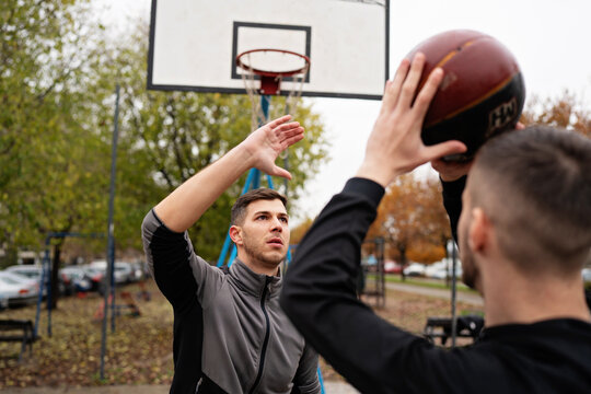 Two athletic friends engaging in a competitive match, with one player defending while the opponent shoots towards the hoop on an autumn park court.