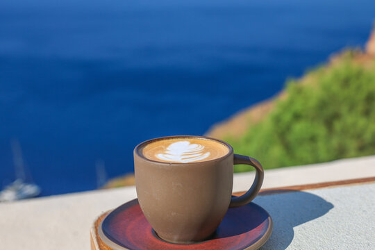 Brown ceramic cup of latte with leaf foam art rests on a ledge overlooking the vivid blue Aegean Sea in Santorini, blending cozy cafe culture with a stunning Mediterranean coastal view.