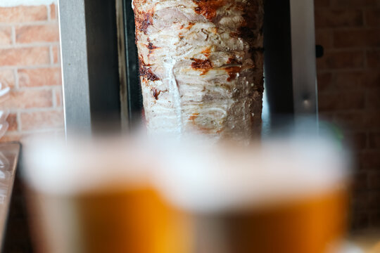 Golden‑brown gyro meat roasts on a vertical grill while blurred beer glasses sit in the foreground, capturing the casual, social atmosphere of a Greek taverna serving Mediterranean street food.
