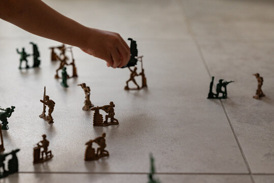 A close-up shot of a child's hand positioning miniature green and brown plastic army men on a tiled floor during a playful war game