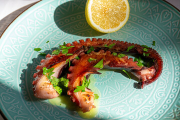Tender grilled octopus tentacles with lemon half and chopped parsley on a turquoise patterned plate © ElenaEdelweys