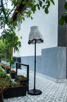 A vertical shot of a tall, black metal standing floor lamp featuring a black and white patterned lampshade, positioned next to a green planter box on an outdoor tiled patio walkway.