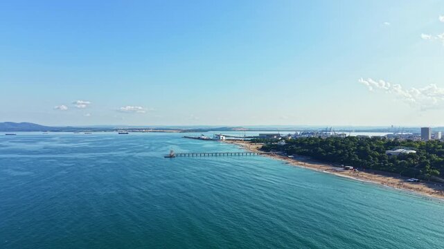 Waves break gently on the sandy beach while boats move in the water. A pier extends into the sea while buildings stand in the background. Cloudless sky adds to the view.