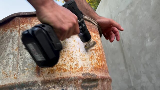 Handyman using a cordless drill driver with a wire brush attachment to remove rust and old paint from a large, dented, and weathered iron barrel outdoors