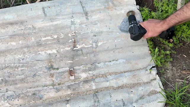 Unrecognizable male worker's hands using a cordless drill equipped with a round wire brush to remove old paint and rust from a weathered, corrugated metal sheet in a backyard