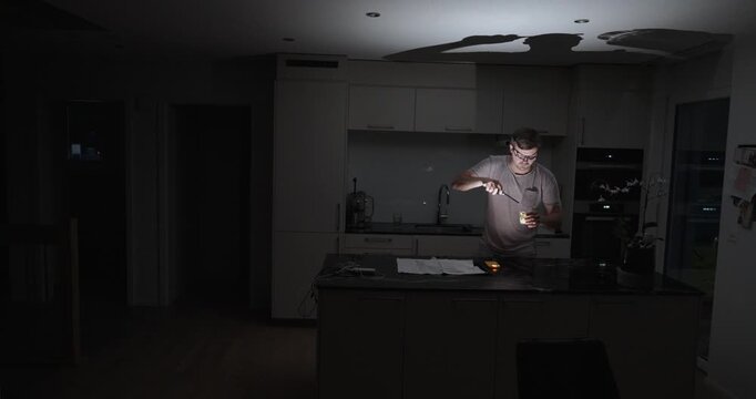 A man searches through a dark kitchen with a flashlight during a power outage, finds a candle, and lights it to illuminate the room. Realistic home emergency and blackout preparedness concept.