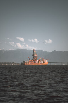 Telephoto view of an offshore drilling vessel on dark coastal water, framed by a long bridge, hazy mountain ridge and scattered clouds beneath an expansive sky