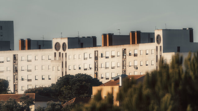 Telephoto view of a weathered multi-story residential block with repetitive shuttered windows, narrow balconies, rooftop chimneys and trees in warm evening light