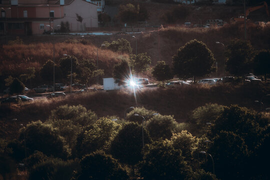 Bright starburst sunlight reflects from a car windshield among parked vehicles, trees and streetlights on a dry suburban hillside in a moody telephoto city scene