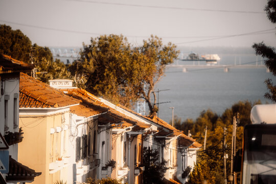 Telephoto view down a sunlit row of tiled residential roofs toward a broad river, distant cargo vessel and bridge, with trees, utility wires and part of a bus