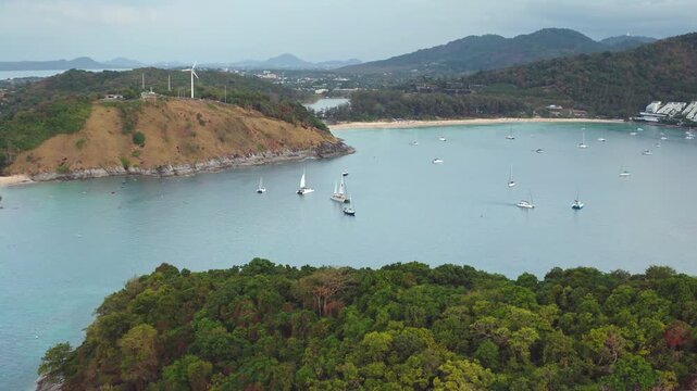 Aerial view of Nai Harn Beach in Phuket, featuring a beautiful tropical bay dotted with sailboats and a prominent wind turbine on the hill. Travel and explore Thailand. Drone flight panorama