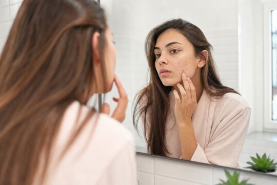 Young woman examining acne on her face in bathroom mirror