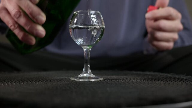 Male hands pouring alcohol into a small glass. Unrecognizable male hands opening a green bottle and carefully pouring a clear alcoholic beverage, like vodka or gin, into a small stemmed glass