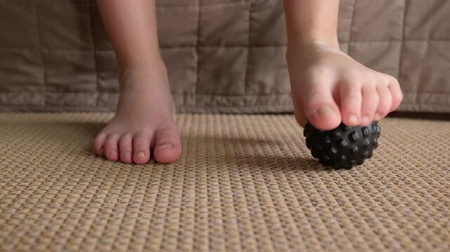 Child massaging foot with spiky ball for flatfoot prevention. Kid's feet doing physical therapy exercises with a black massage ball on the floor, preventing flatfoot and improving muscle tone