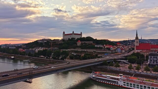 Castle overlooks the city and river during sunset. Boats float in the water while cars drive on the bridge. Buildings line the streets under a colorful sky.