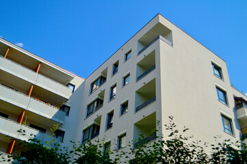 Fototapeta premium Modern residential apartment building featuring sleek white minimalist architecture with balconies and green foliage under a vibrant blue sky.