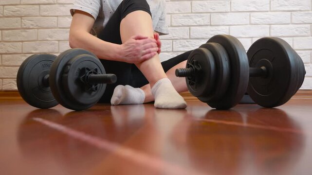 Suffering leg pain after dumbbell workout at home. Woman feeling intense pain in her leg and massaging her shin after a strenuous workout with dumbbells, highlighting the risks of sports injuries