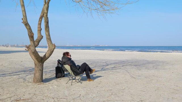Woman relaxing on a folding chair at the winter beach. Solitary woman in a winter jacket sitting on a portable chair under a bare tree, peacefully watching the sea on a deserted sandy beach