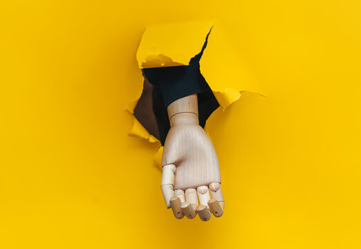 A wooden hand protrudes through a torn hole in yellow paper, begging for alms. Concepts of prosthetics, disability, and poverty.