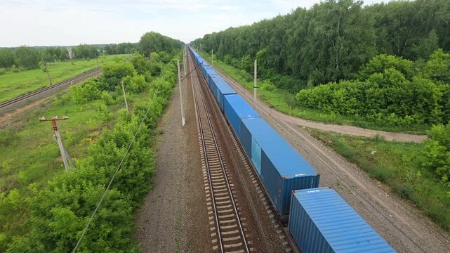 High angle view of a long freight train carrying blue shipping containers moving along a railway track through a rural green forest landscape on an overcast day